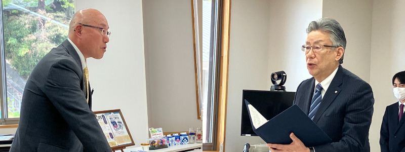 室内でスーツ姿の男性2人が向かい合い、右の男性が書類を手に読み上げている様子。背景には窓やパソコンがあり、後方にマスクを着けた人物も立っている。