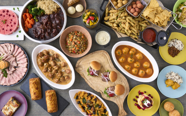 テーブルいっぱいに世界各国の料理が並ぶ俯瞰写真。ビーフシチューやミートボール、パスタ、スライダー、揚げ物、デリサラダ、点心、チップス、ハムの盛り合わせ、デザート（ケーキやパンケーキ）など、多彩なメニューがカラフルな器に盛り付けられている。