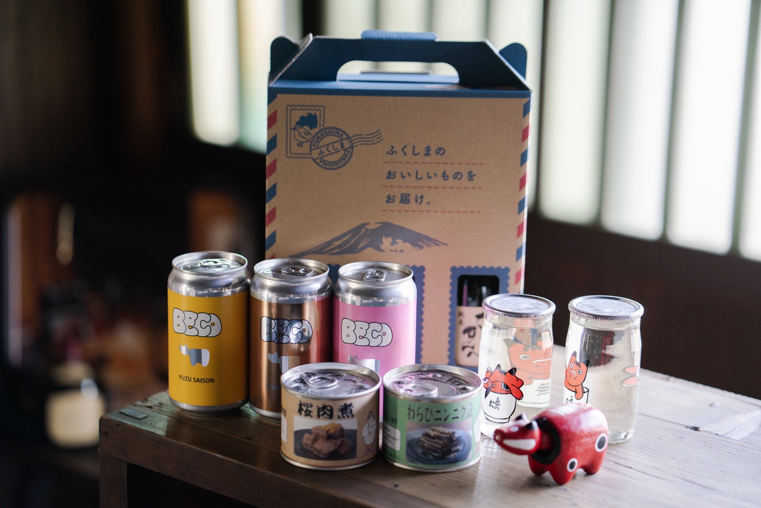 五十嵐太右ヱ門:福島の特産品を詰め合わせたギフトセット。クラフトビールの缶、日本酒の瓶、缶詰、赤べこが木のテーブルに並び、背後に配送用ボックスが置かれている。